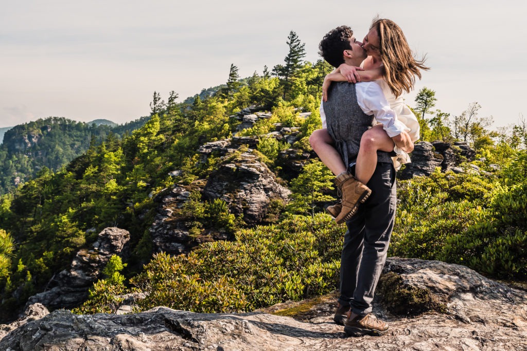 A bride in a short wedding dress jumps up and wraps her legs around her husband as they share a passionate kiss on a rugged mountain overlook.