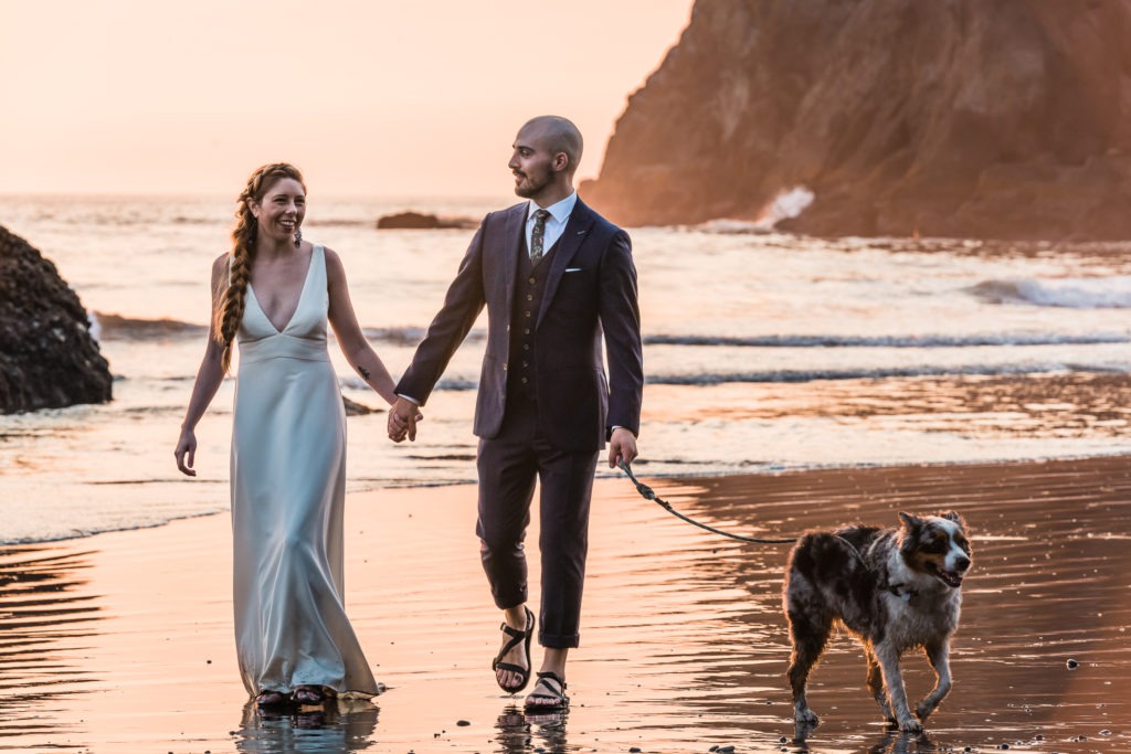 At this wedding with their dog, Elena, Parker and their pup, Winter, stroll down the beach at sunset, the water lapping at their feet and shades of pink glistening in the waves.