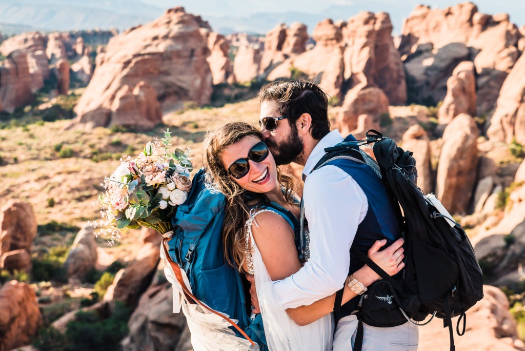 Moab elopement wedding, Utah. The bride, with her bouquet in her backpack and her sunglasses on, smiles at the camera as her new husband pulls her in for a kiss, with wacky red rock formations rising up in the background.