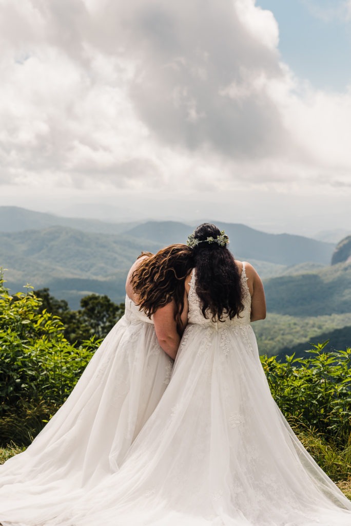 After their same-sex wedding, two brides in flowing white dresses cuddle close together and look out over a lush green forest in North Carolina.