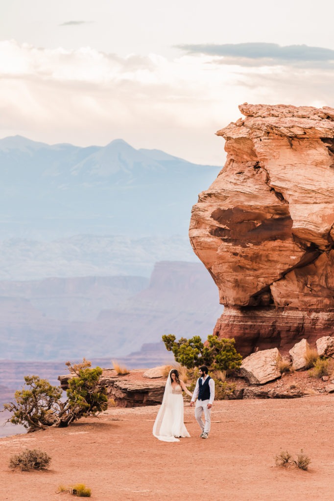 Moab wedding, Utah. The couple walk hand in hand towards the camera, with a huge red rock formation rising up behind.