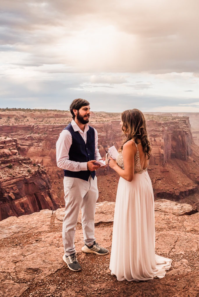 Sunset wedding ceremony in Moab, Utah. The couple stand on the edge or a dramatic red canyon to share their vows.