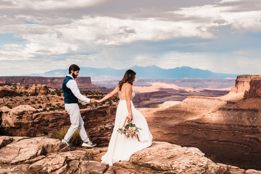 Moab wedding. The bride in a flowy white dress leads her groom by the hand across the red rocks, with a huge canyon leading towards mountains in the distance.