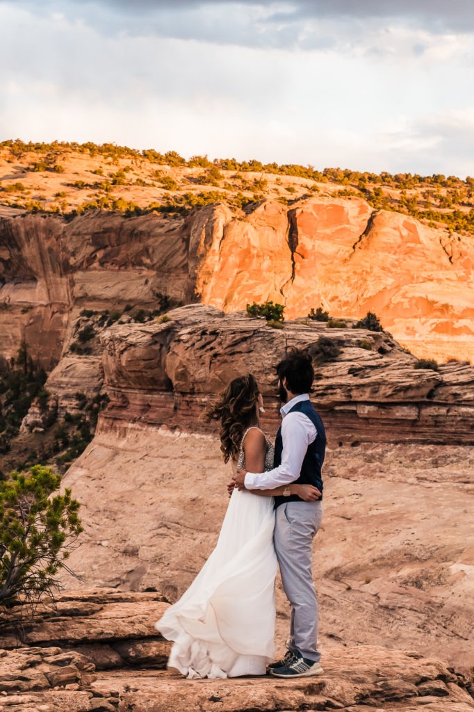 The bride's flowy dress catches the wind as the couple cuddle up and watch the red rocks of Moab light up at sunset.