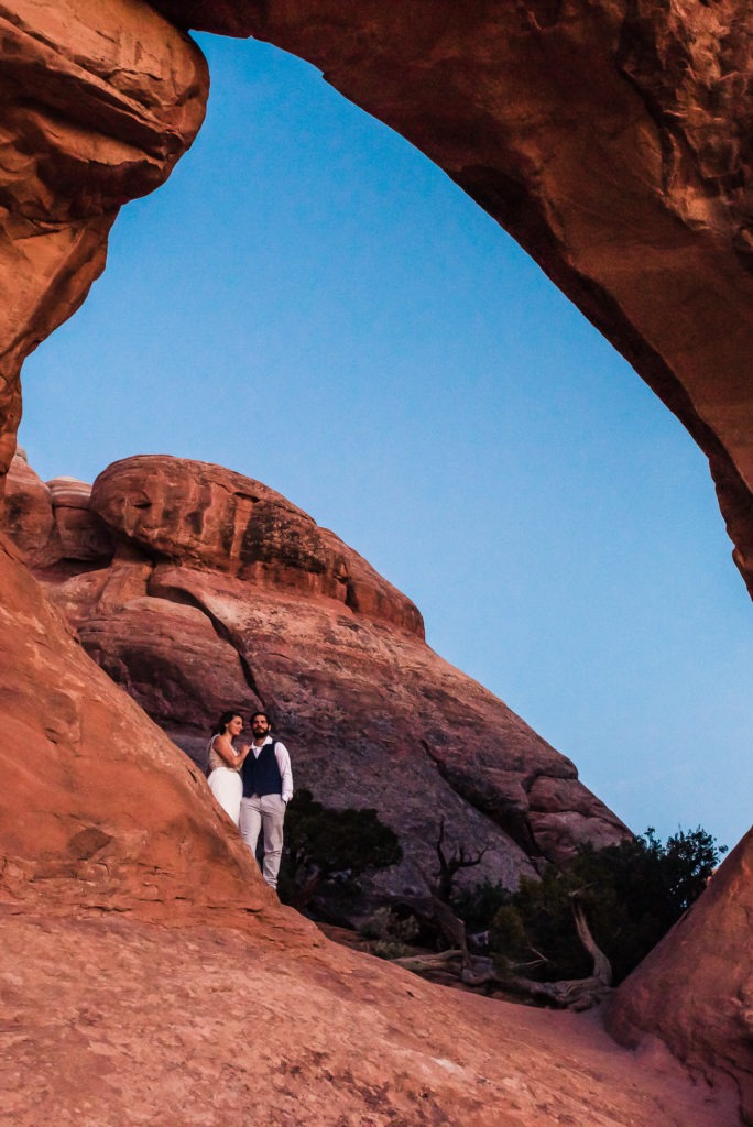 Sunrise wedding, Moab. The soon-to-be-married couple stand below a red archway and watch the rocks turn a deep red color as the sun rises and hits them straight on.
