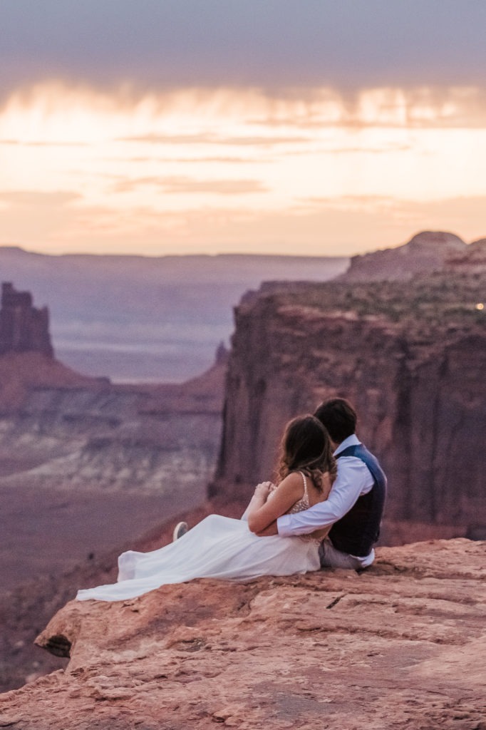Sunset Moab wedding. The couple sit cuddled up on the edge of the Canyonlands, watching the sky turn golden at sunset.