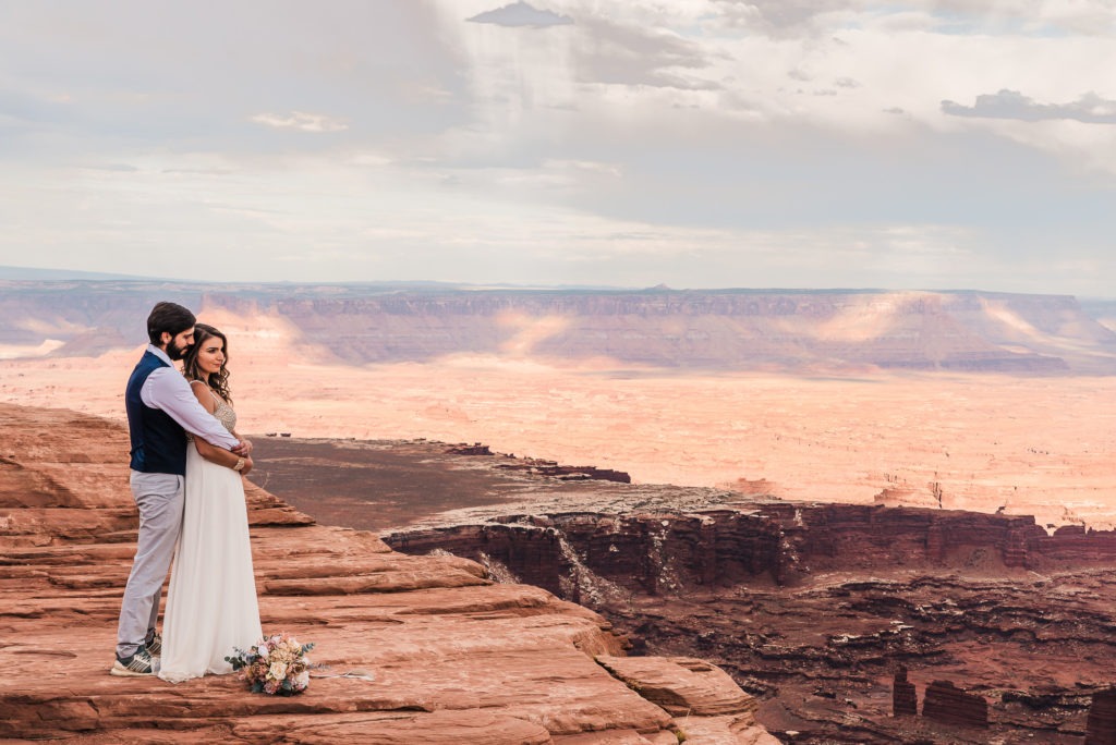 Moab adventure elopement. The groom cuddles his bride from behind as they stand on the edge of a canyon and take in the views.