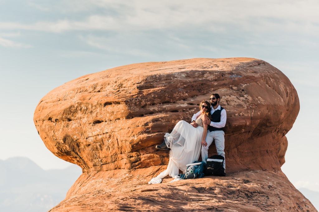 At their Arches National Park wedding, the couple snuggle up on a huge red rock formation, sunglasses on and backpacks at their feet. They take a bit of time out to relax and enjoy the views.