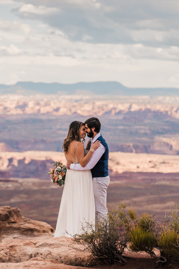 Moab elopement. The couple snuggle up on the edge of a huge canyon, with endless views behind them.
