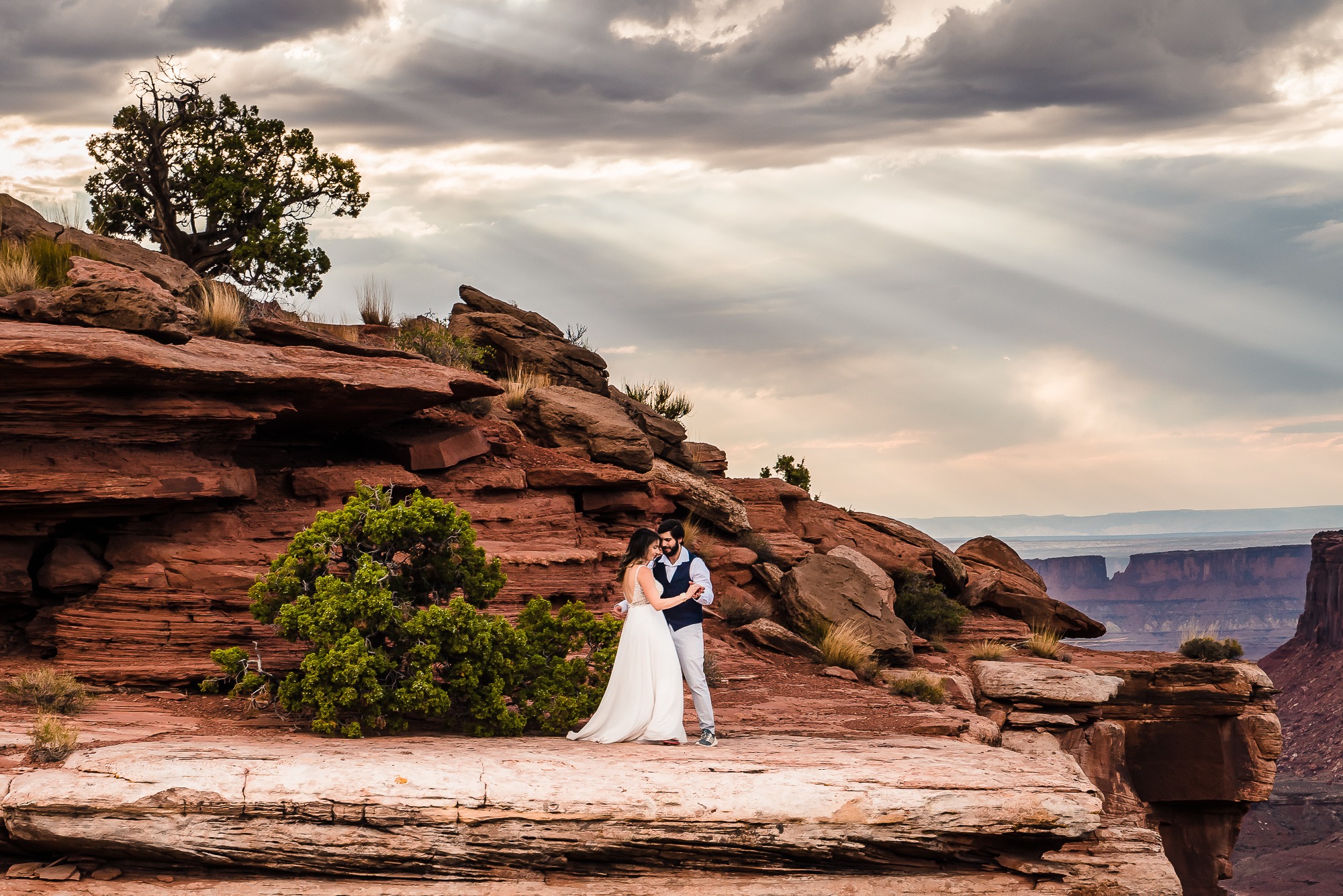 To celebrate their Moab elopement, this couple share a first dance on a dramatic overlook, with moody, cloudy skies above and rays of sunshine pouring down on them.