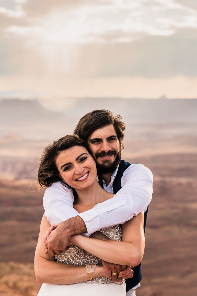 Moab, Utah elopement wedding. A handsome man, with dark hair and a dark beard, wraps his arms around his wife from behind as they cuddle up and smile straight at the camera. The rugged canyonlands of the Moab region extend behind them and a beautiful golden light is pushing through the clouds.