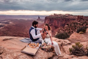 As the sun sets on their Moab elopement, this couple sit together on the rocks and share a pizza and beer for their wedding celebrations. There are towering red rock ‘hoodoos’ in the distance and a dark, dramatic sky above.