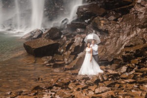 Waterfall elopement. The couple take a clear umbrella and venture to the very edge of the waterfall.