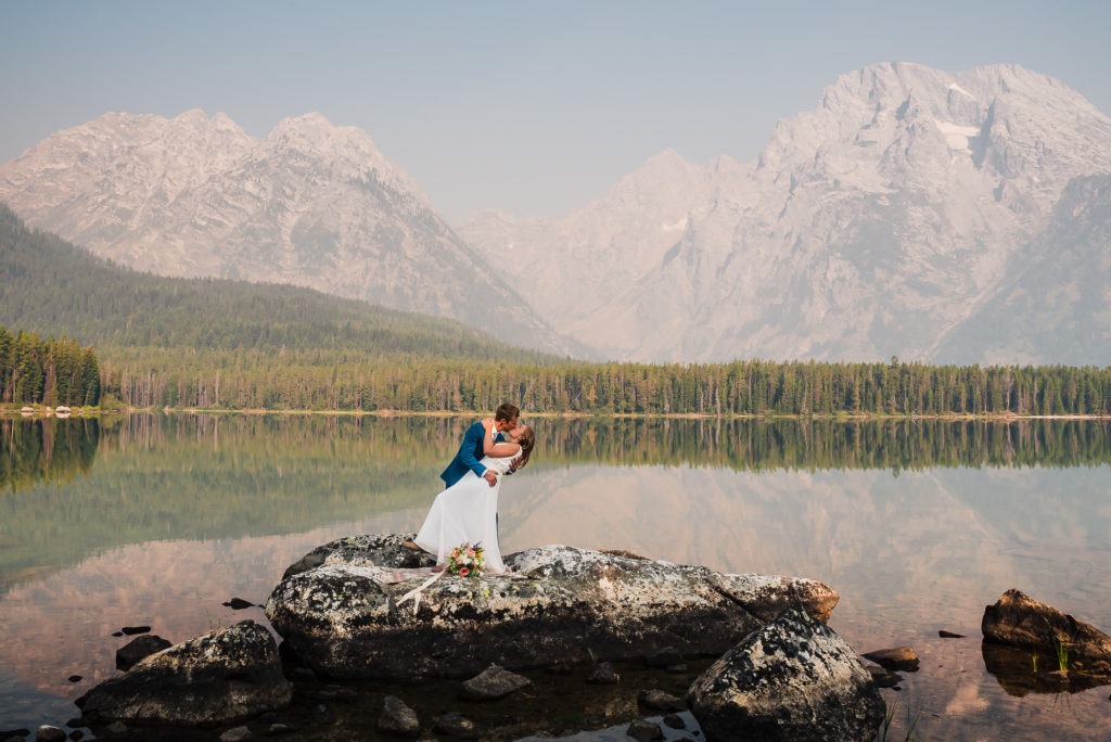 The groom dips his wife for a kiss on the edge of a lake with huge mountains rising in the background.