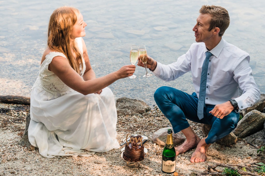 A couple in their wedding clothes sit by the edge of a lake and enjoy champagne and chocolate cake to celebrate.