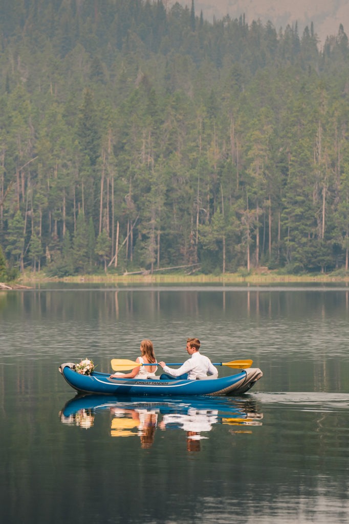 Adventure wedding. A couple are rowing a kayak across a calm lake, their image perfectly reflected in the water.