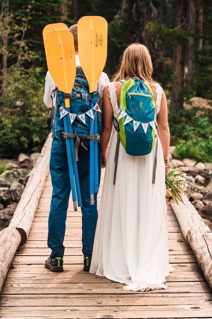 Just married signs hang from their backpacks as the couple walk hand in hand over a wooden bridge.