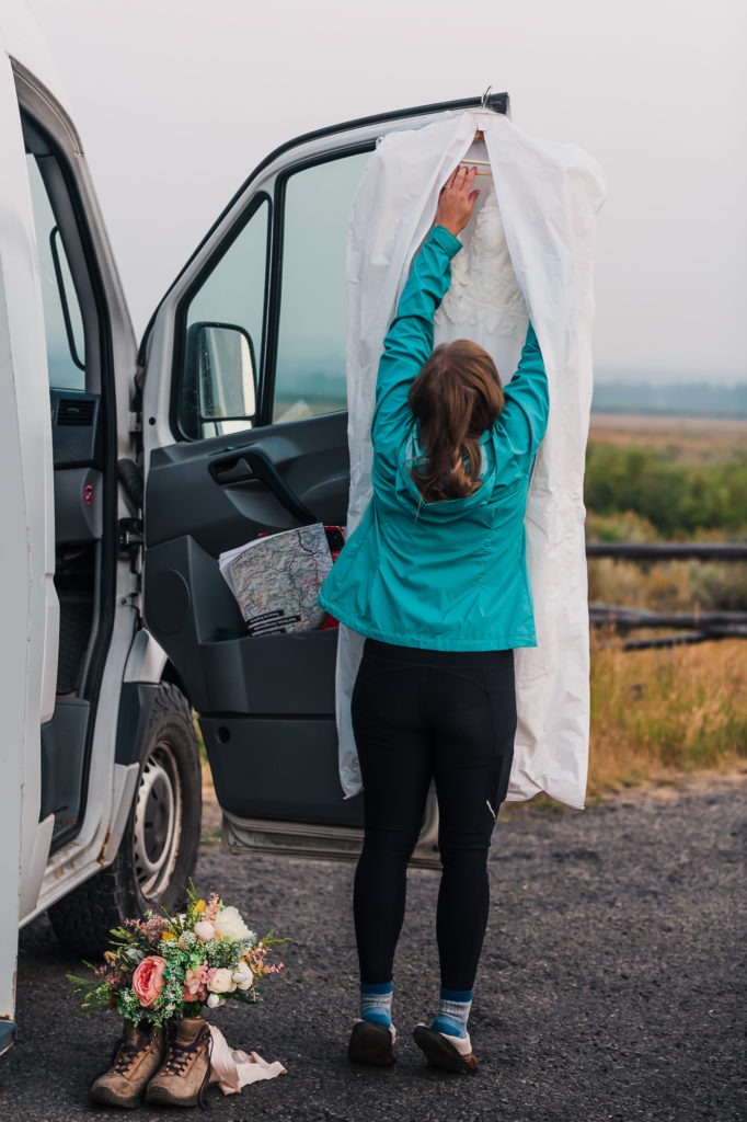 A woman's wedding dress is hanging from the door of her van and she is reaching up to get it. Her hiking boots and bouquet are to the side.