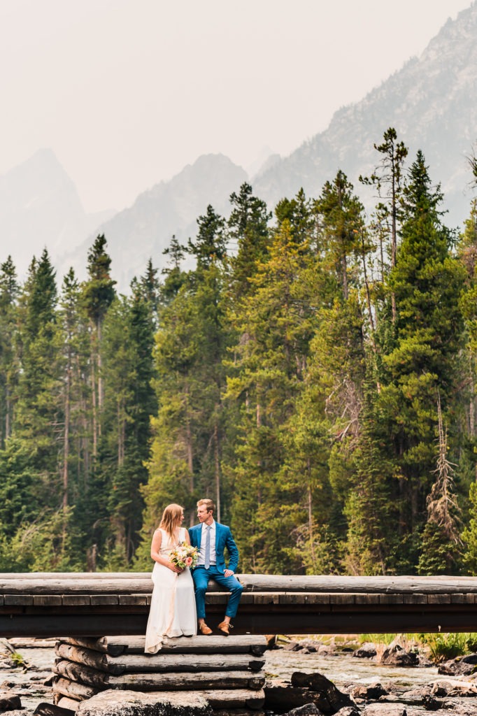 A couple site on a wooden bridge with tall trees rising up behind them and mountains behind that.