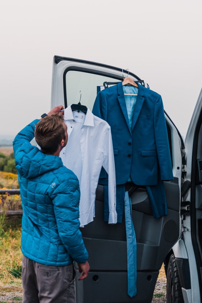 A man's wedding attire is hanging from the door of his van and he is lifting down his shirt to start getting ready.