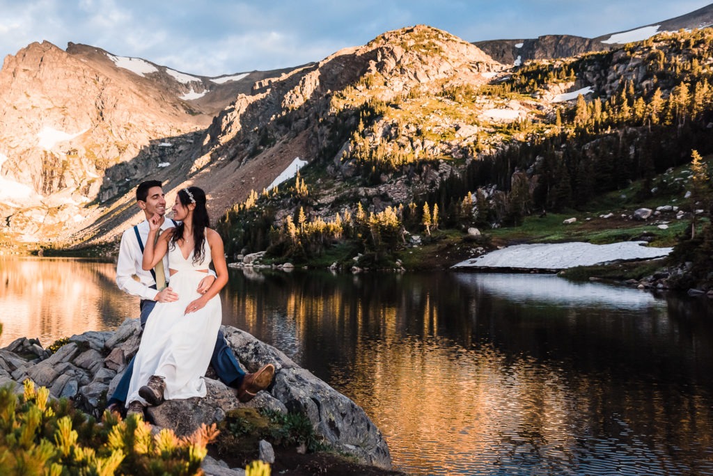 Sunrise wedding. At their Colorado elopement, the couple sit by an alpine lake and enjoy the sunrise which is casting a golden glow over the mountains.
