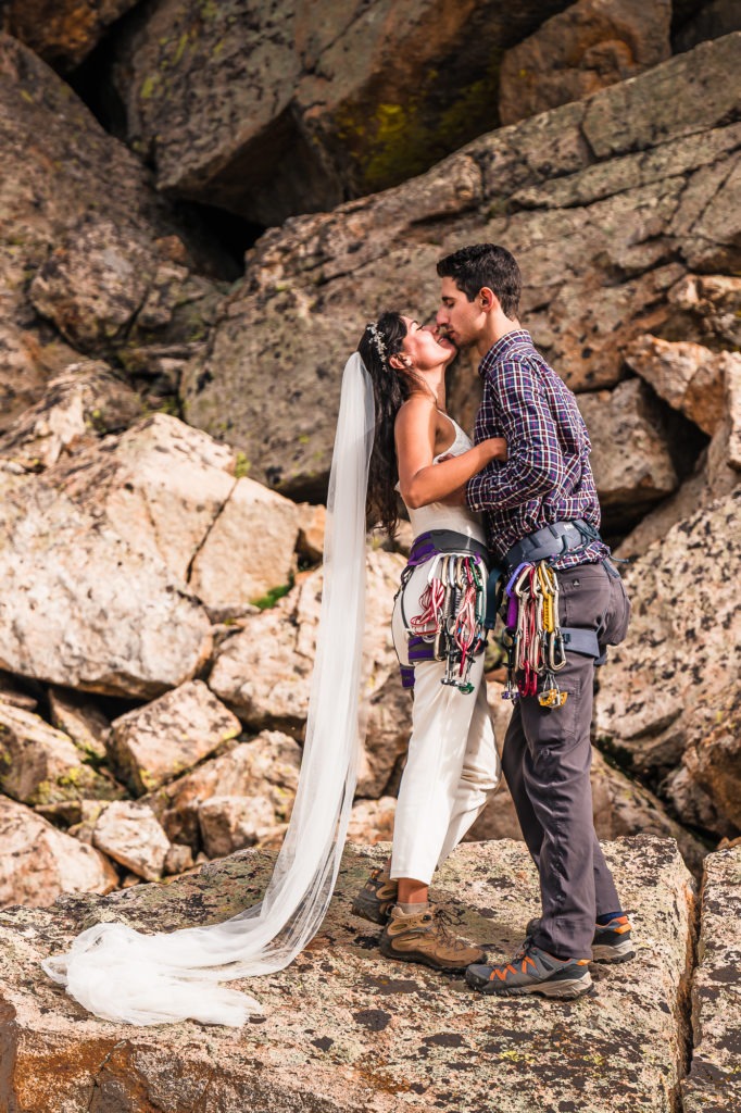 Rock climbing wedding. The couple share a kiss in their wedding and climbing gear. She has on a white jump suit and a long white veil that flows behind her.