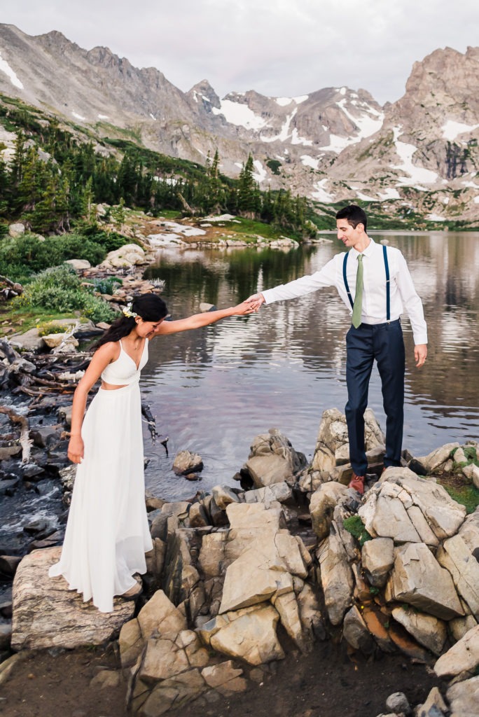 Hiking wedding. The groom helps his bride over the rocks by the edge of a mountain lake.