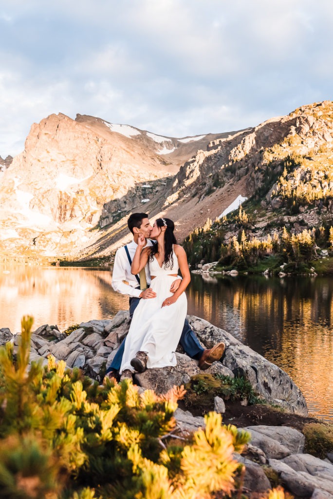 Sunrise wedding. The couple snuggle on a rock by an alpine lake as the rocky mountains behind them light up in the early morning glow.
