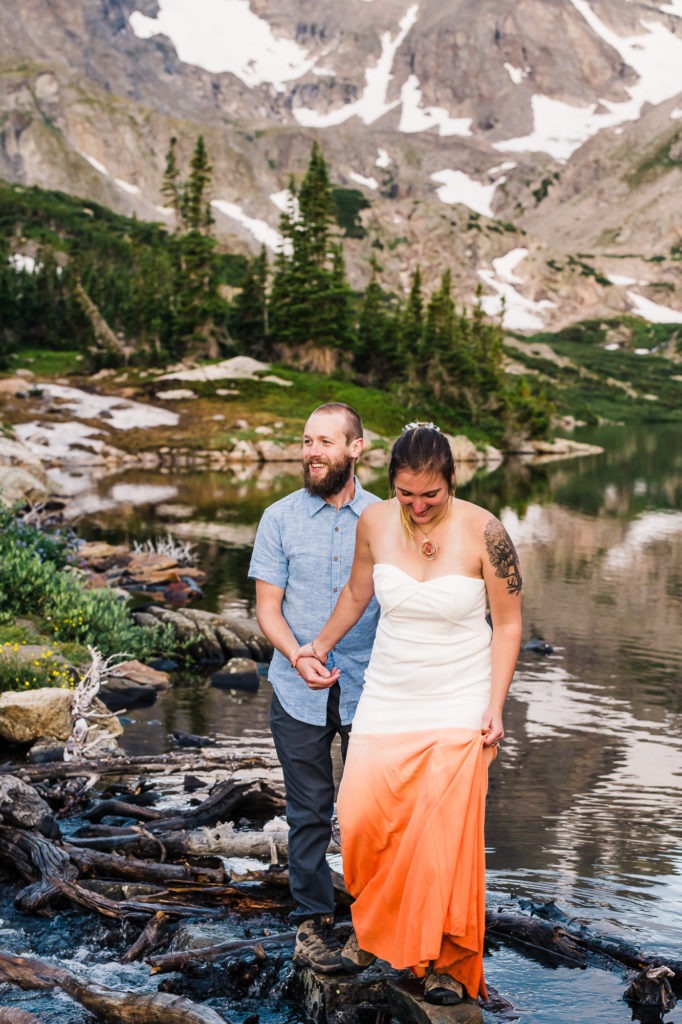 A young couple explore around an alpine lake for their Colorado mountain engagement session. She wears a white dress with a dip-dye orange bottom half.