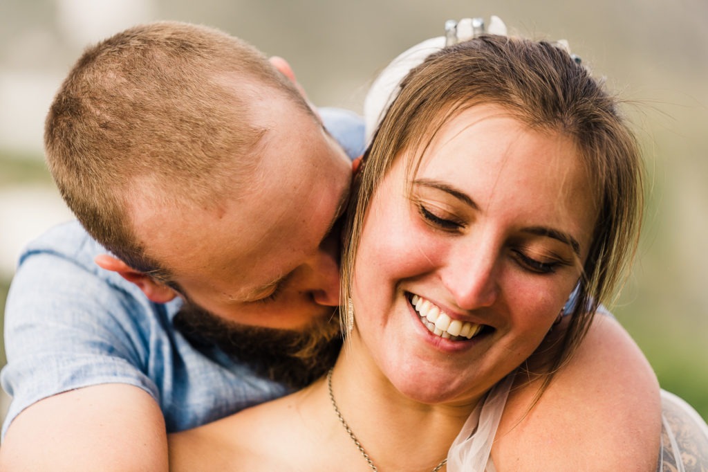 The bride-to-be closes her eyes and smiles as her fiance cuddles her from behind and kisses her neck