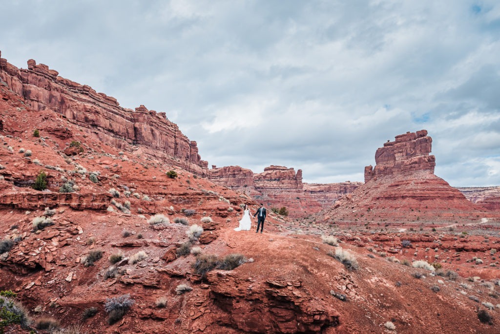 Hiking wedding in Moab, Utah. The couple hold hands in the middle of an enormous red landscape.