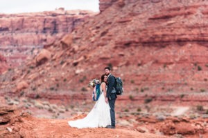 Adventure elopement, Moab. A couple with backpacks on cuddle up, with towering red rocks behind them. The bride has a bouquet in her backpack.
