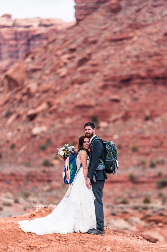 Adventure elopement in Moab, Utah. A bride and groom both with backpacks on cuddle up face to face with red rocks in the background.