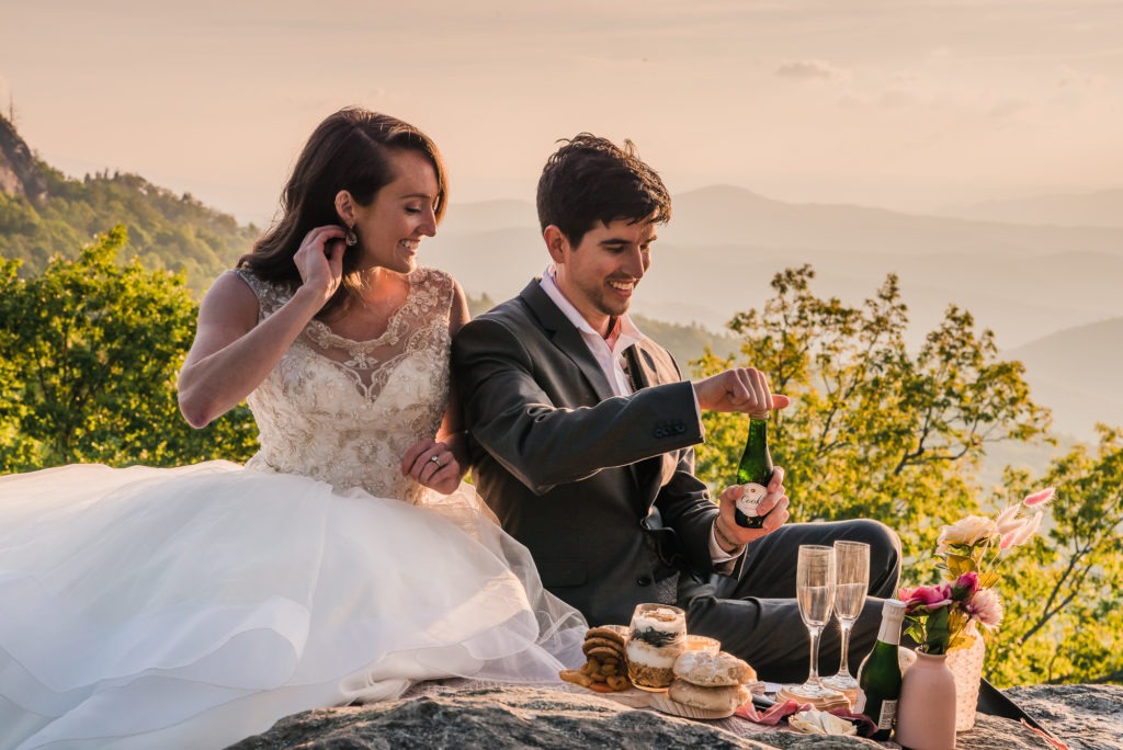 The couple share a champagne picnic for breakfast on a rocky overlook with mountains in the background.