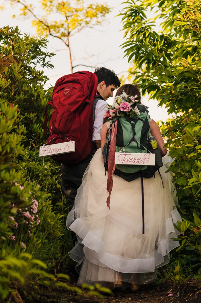 The couple have their wedding clothes on and backpacks with signs reading 'Always and Forever'.