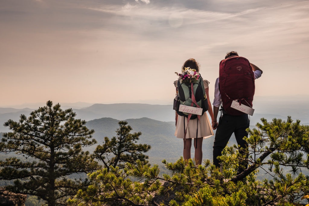 During their hiking anniversary trip, the couple stand side by side, backpacks on, and look out over the distant mountain ranges.