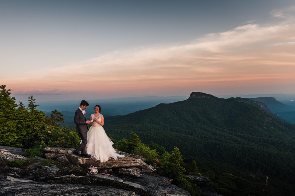 The couple re-share their wedding vows during their anniversary session. They stand on a grey rock atop a mountain, with the sun setting behind them.