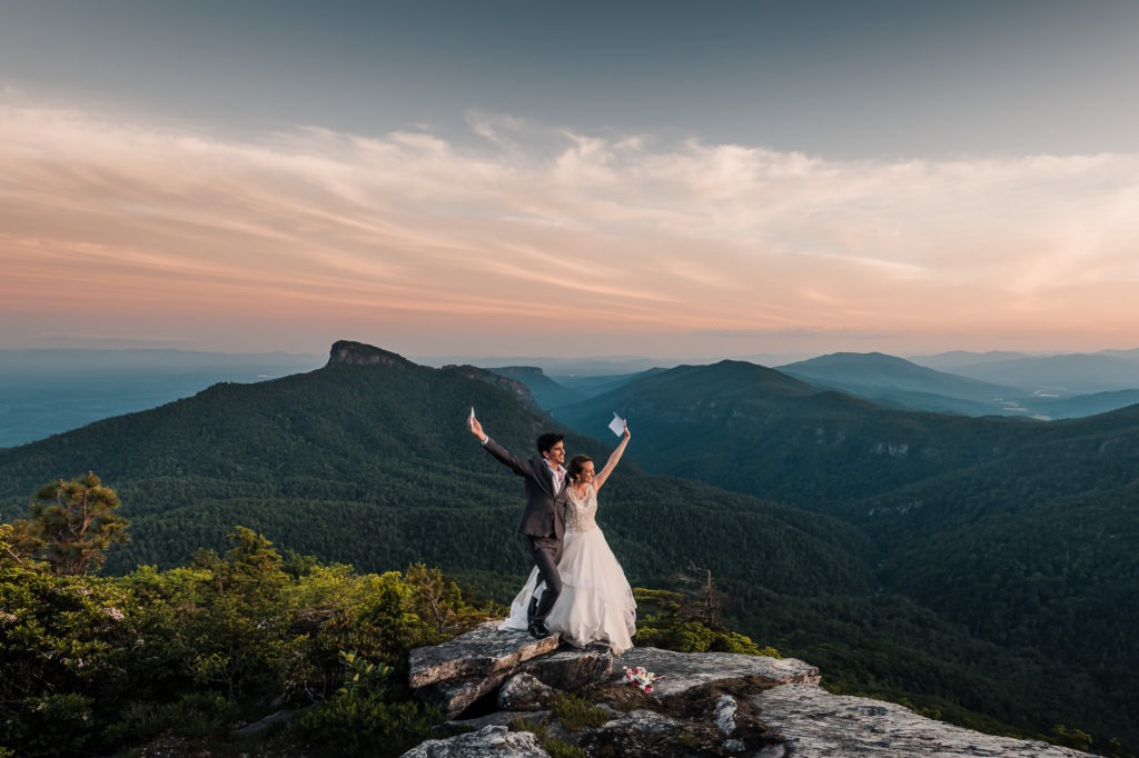 After sharing their vows, the couple raise their arms in the air and celebrate! The sky is turning pink over the mountains behind them.