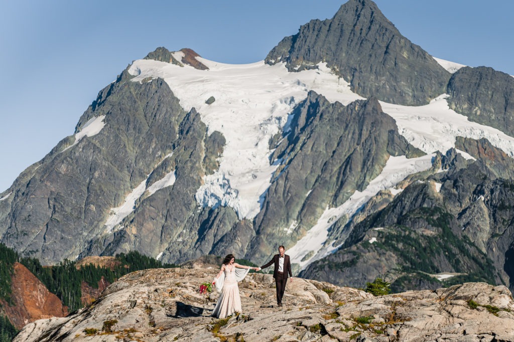 North Cascades, Washington, mountain elopement. The couple walks hand in hand towards the camera, with an enormous snowy peak rising behind them.