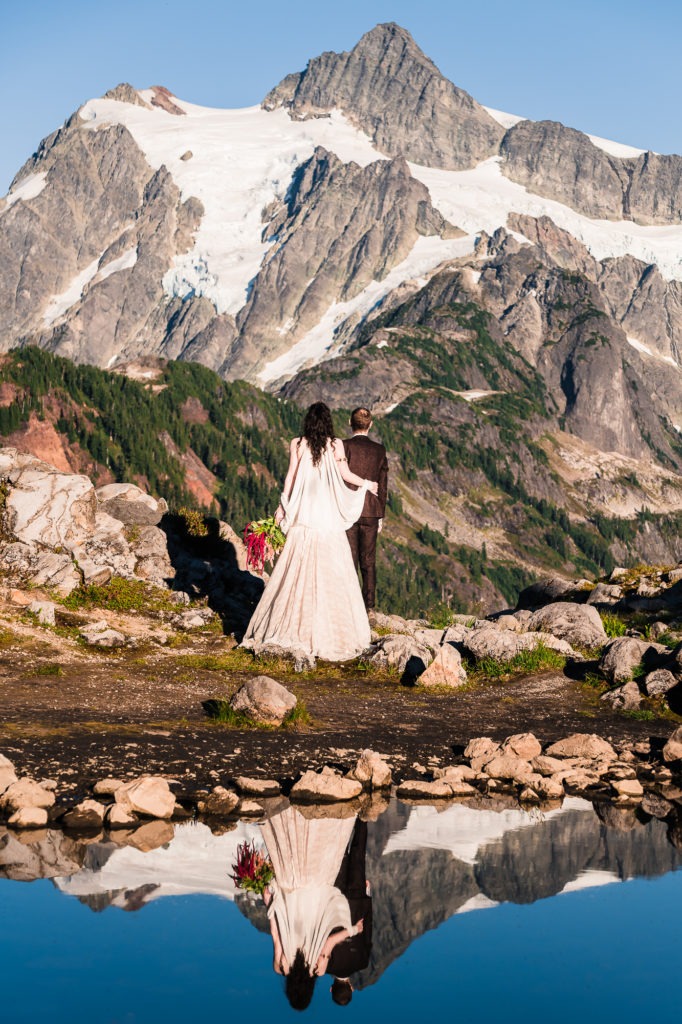 Mountain elopement. The couple are side by side looking out to a dramatic snow-capped peak. Their is a small pond behind them and their reflection is cast perfectly in the still water.