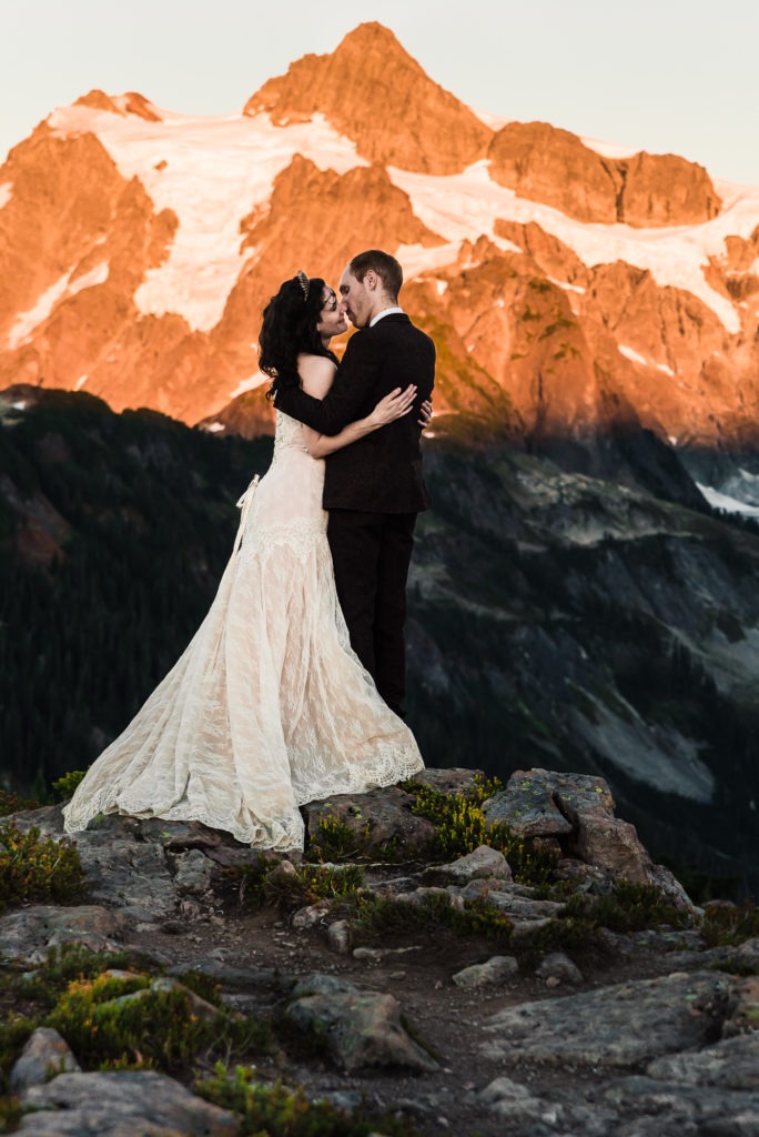 Mountain sunset elopement. The couple share a kiss as the setting sun casts an 'alpen glow' on the towering mountain behind, turning the rocks and snow a beautiful pinky golden hue.