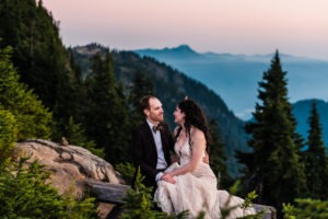 North Cascades, Washington elopement. The couple cuddle together sitting on a wooden bench overlooking lush forest and misty mountains. They're a striking, alternative couple: he is in a brown suit and metallic bow tie, she has a crystal crown and jewelled headpiece.