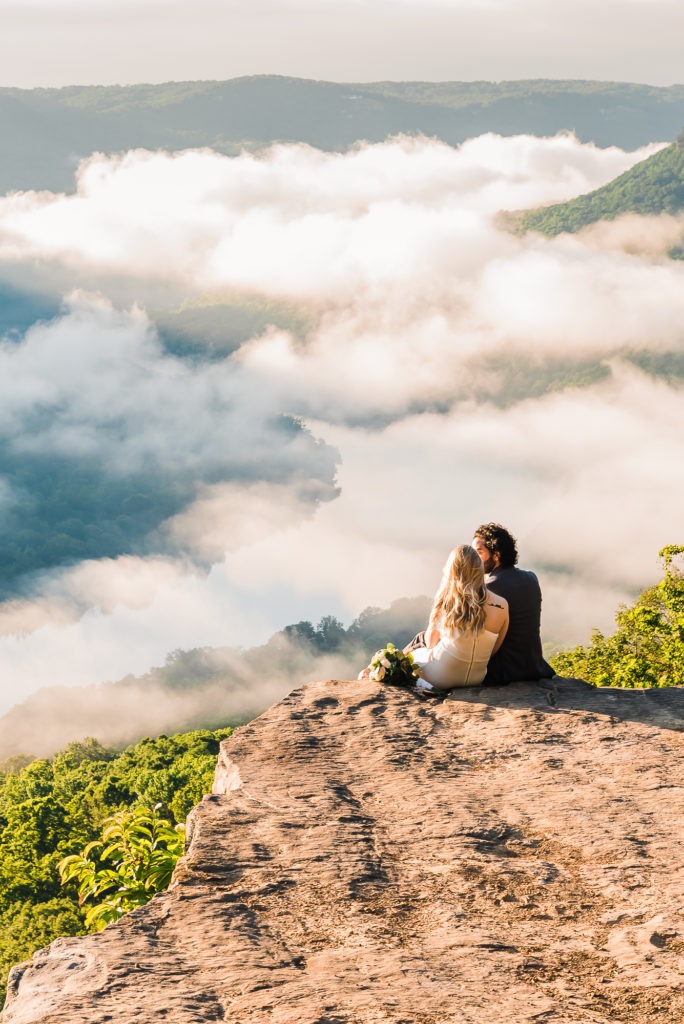 Sunrise elopement, Tennessee. The couple sit side by side on a rocky ledge, overlooking a winding river below. Morning mist fills the valley and they look to be floating on clouds.