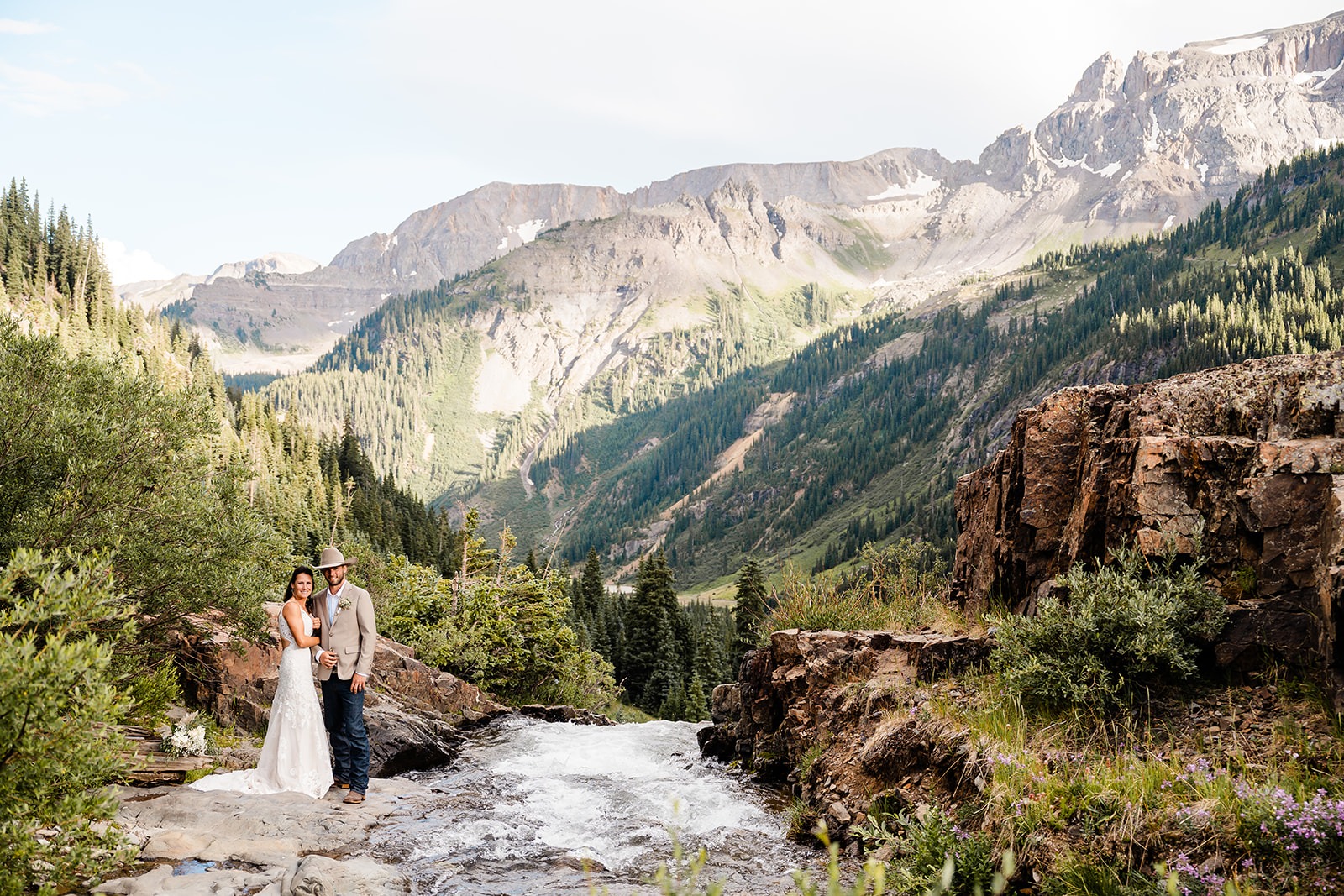 A wedding couple standing close together on the side of a river in the mountains
