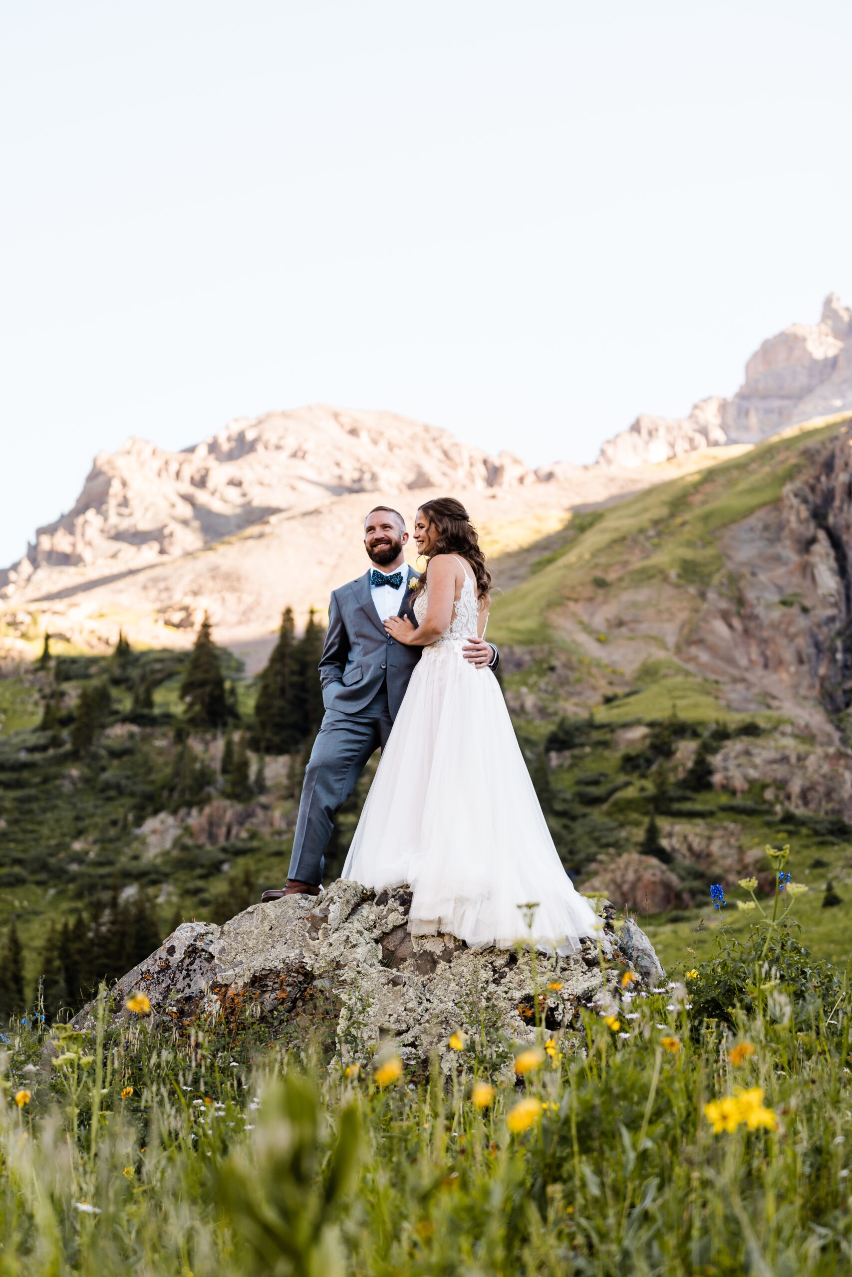 A newlywed couple standing on a rocky in a grassy valley in the mountains smiling with their arms around each other