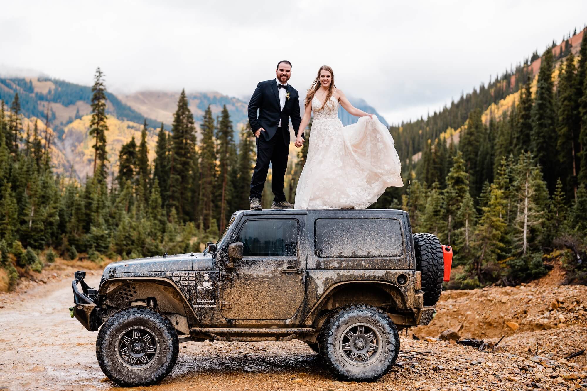 A newlywed couple standing on top of a Jeep smiling together in the mountains