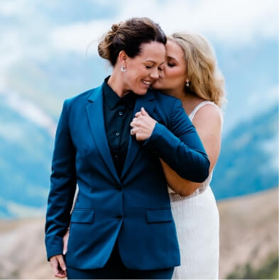 A person in a dress leans in to kiss the cheek of someone in a tuxedo from behind.