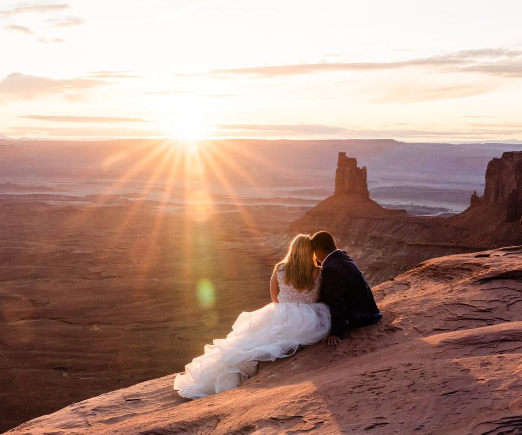 A couple leans into one another, head to head, looking out as the sun sets over the Utah desert.