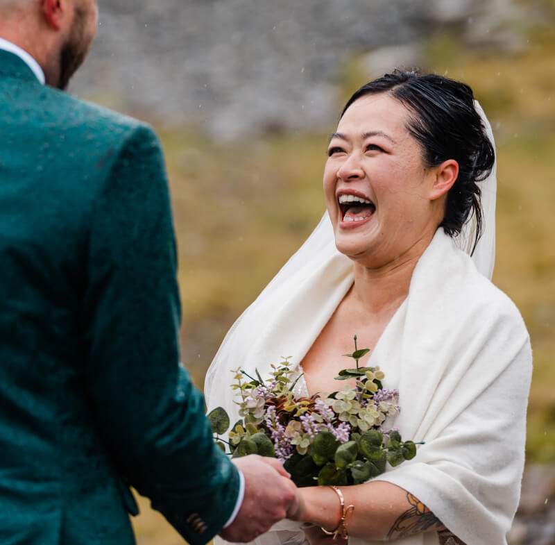 A person in a wedding dress smiles cheerfully holding their partner's hands.