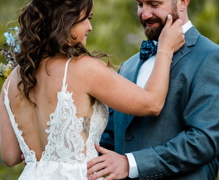 A person in a dress leans in to touch the face of someone in a tuxedo, both smiling at each other.